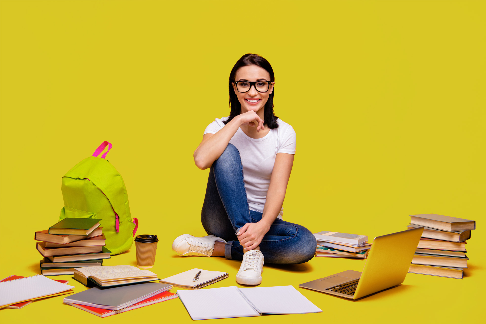 A smiling graduate student sits cross-legged on the floor surrounded by study materials including a laptop, notebooks, coffee, and textbooks. She wears glasses, a white t-shirt, and jeans, symbolizing a modern, confident learner balancing education with life. The vibrant yellow background reflects the energy and optimism of pursuing a flexible, career-focused graduate degree at Our Lady of the Lake University (OLLU).